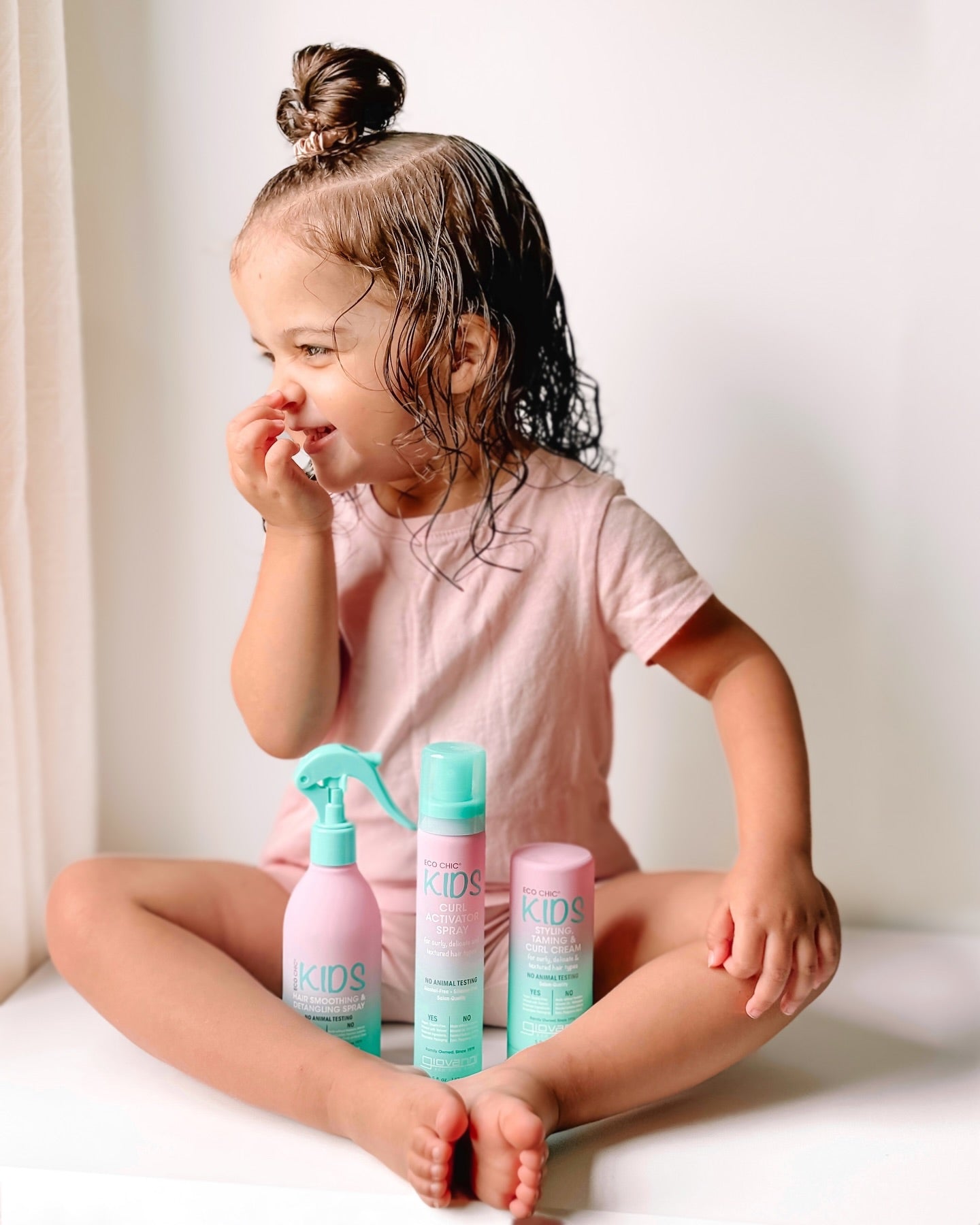 A young child with wet hair, smiling in a pink outfit, sits cross-legged with three turquoise and pink bottles of Giovanni Cosmetics ECO CHIC® KIDS CURL ACTIVATOR SPRAY for moisture retention placed in front of them.