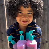 A young child with curly hair smiles while holding several bottles of Giovanni Cosmetics ECO CHIC® KIDS 2 IN 1 SHAMPOO & CONDITIONER, featuring purple bottles with turquoise pumps, standing in sunlight by a wooden fence.