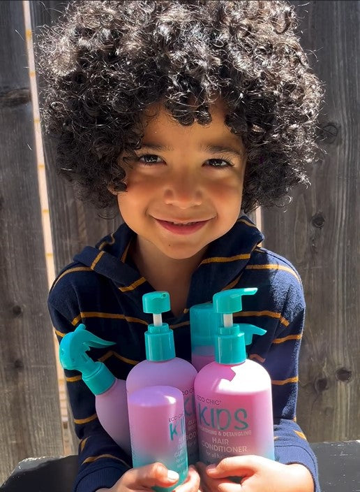 A young child with curly hair smiles while holding several bottles of Giovanni Cosmetics ECO CHIC® KIDS 2 IN 1 SHAMPOO & CONDITIONER, featuring purple bottles with turquoise pumps, standing in sunlight by a wooden fence.
