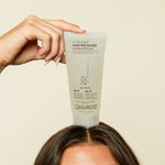 A hand holds a tube of Giovanni Cosmetics MORE BODY™ HAIR THICKENER above a person’s head, the tube resting on their hair against a plain beige background.