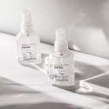 Two clear spray bottles of Giovanni Cosmetics SHINE OF THE TIMES™ HIGH GLOSS HAIR MIST rest on a white surface, illuminated by natural light with soft shadows.