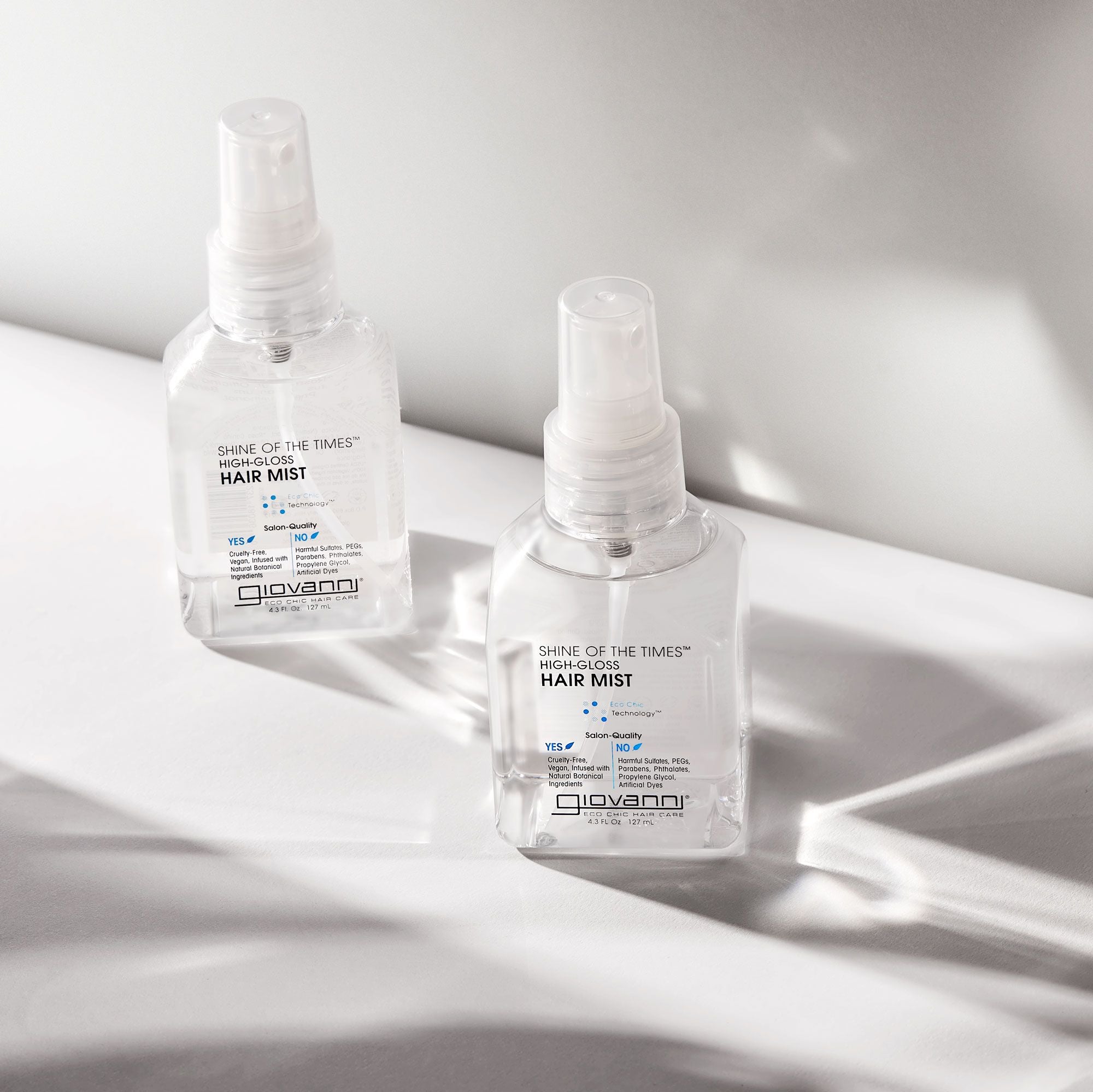 Two clear spray bottles of Giovanni Cosmetics SHINE OF THE TIMES™ HIGH GLOSS HAIR MIST rest on a white surface, illuminated by natural light with soft shadows.