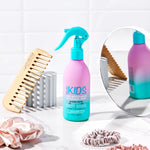A pink and teal bottle of Giovanni Cosmetics ECO CHIC® KIDS HAIR SMOOTHING & DETANGLING SPRAY sits on a white counter with a wooden comb, pink scrunchies, and a round mirror, ready to gently detangle kids' hair by white tiled walls.
