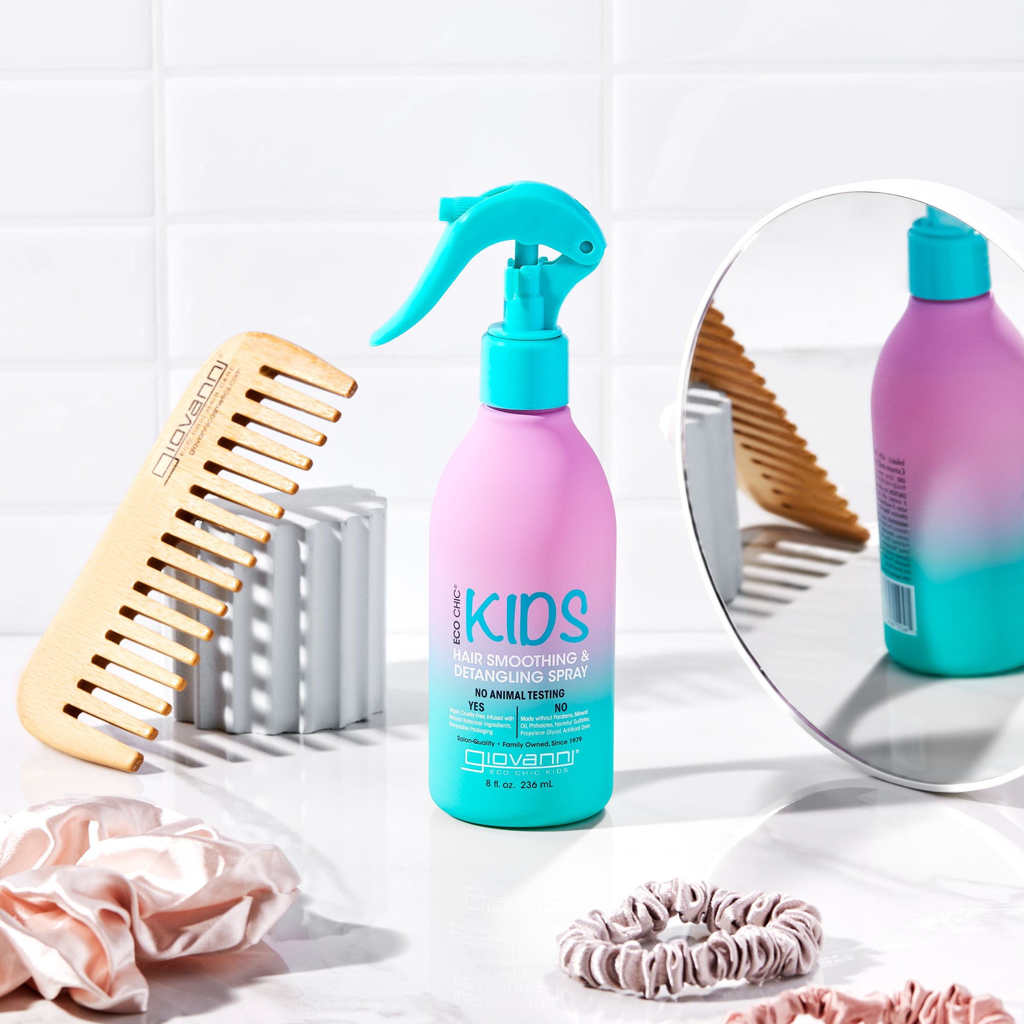 A pink and teal bottle of Giovanni Cosmetics ECO CHIC® KIDS HAIR SMOOTHING & DETANGLING SPRAY sits on a white counter with a wooden comb, pink scrunchies, and a round mirror, ready to gently detangle kids' hair by white tiled walls.