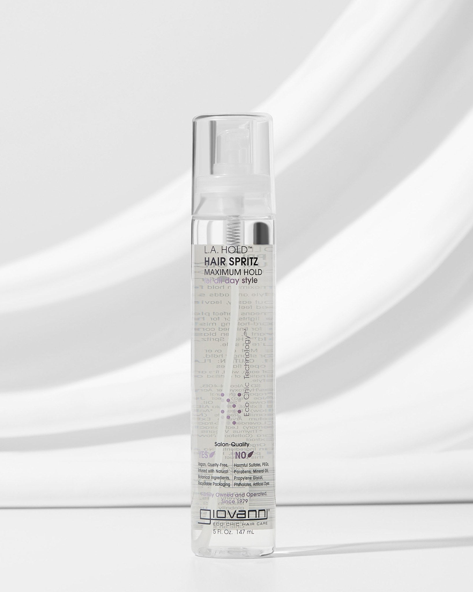 A clear bottle of Giovanni Cosmetics L.A. HOLD™ HAIR SPRITZ stands upright on a white surface against a soft gray background, displaying its visible label and color-safe, salon-quality hair spray inside.