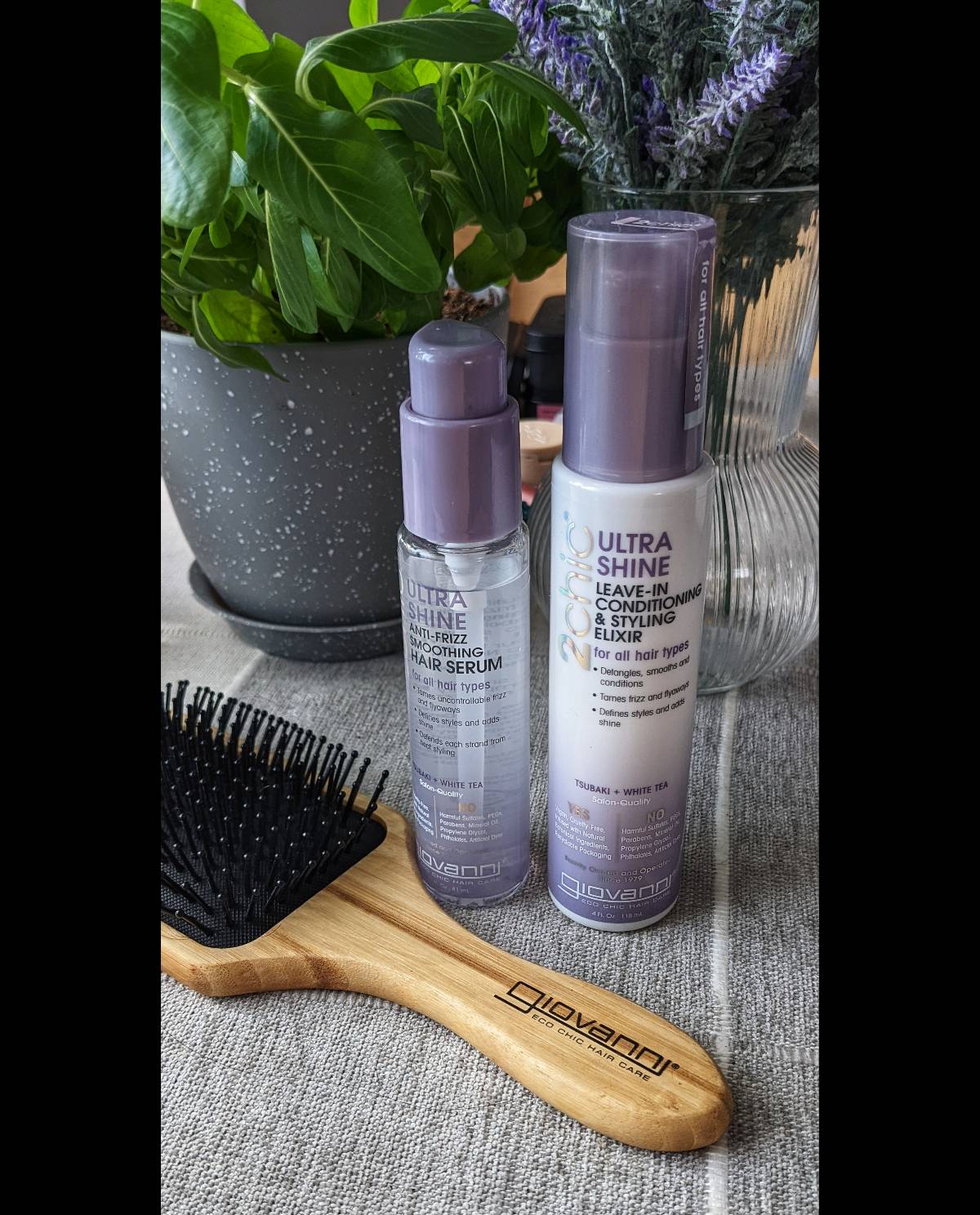 A Giovanni Cosmetics 2chic® ULTRA-SHINE LEAVE-IN CONDITIONING ELIXIR and a wooden hairbrush rest on a table, framed by green plants and a glass vase—a fresh setting inspired by Tsubaki blossom and White Tea.