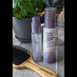 A Giovanni Cosmetics 2chic® ULTRA-SHINE LEAVE-IN CONDITIONING ELIXIR and a wooden hairbrush rest on a table, framed by green plants and a glass vase—a fresh setting inspired by Tsubaki blossom and White Tea.