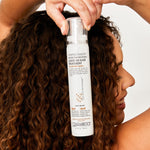A woman with curly brown hair holds a bottle of Giovanni Cosmetics VITAPRO FUSION™ Protective Moisture Leave-In Hair Treatment, highlighting her defined, healthy curls and the color-safe benefits of this hair care product.